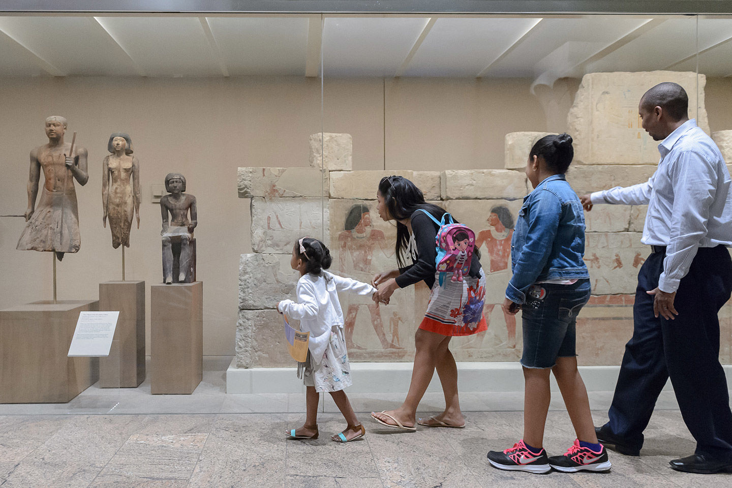A young girl leads her family across a granite-floored gallery of Egyptian stone figures and painted friezes